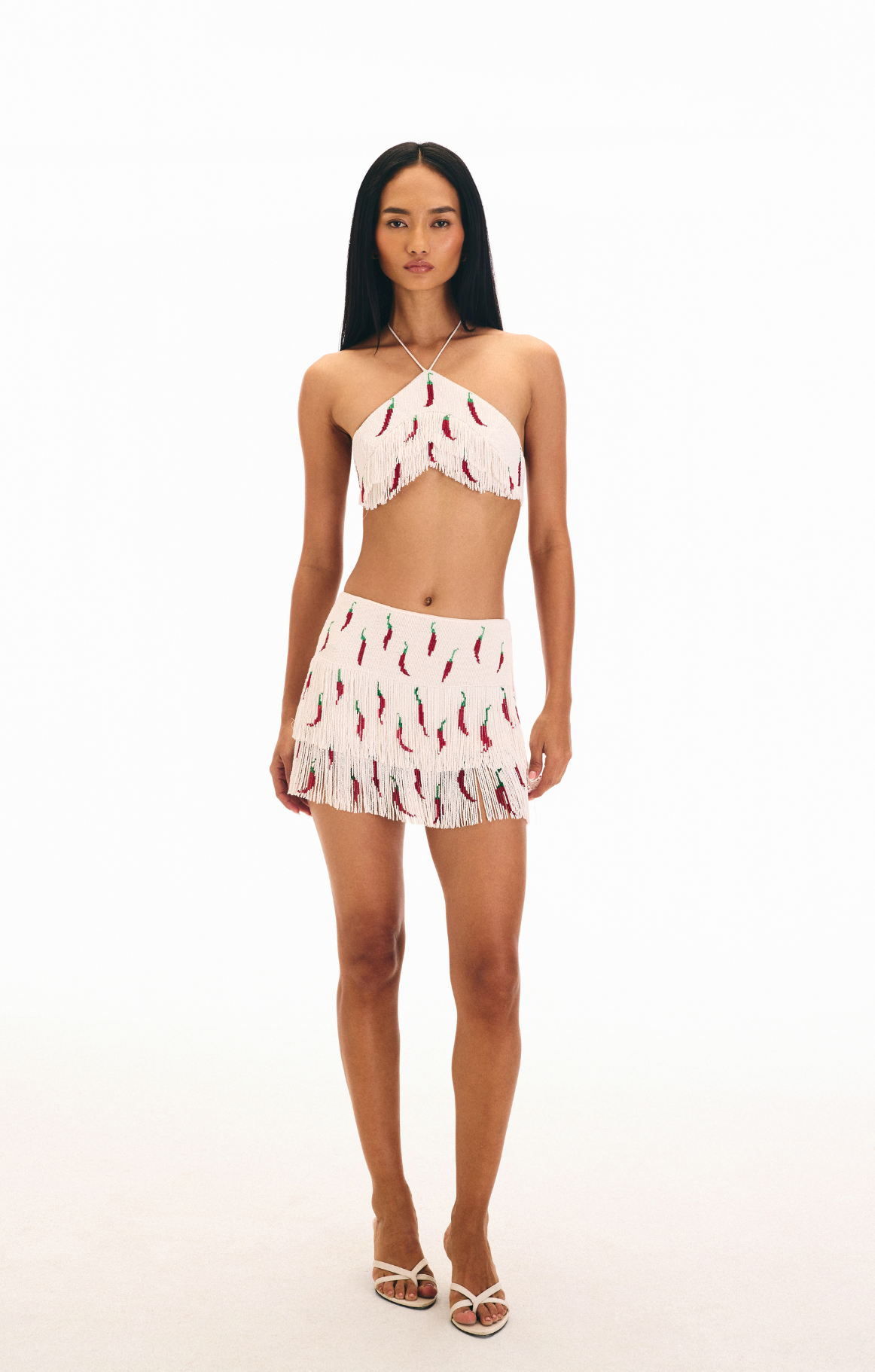 Woman wearing a white cropped triangular halter top and skirt with chili pepper patterns on white tassels. 